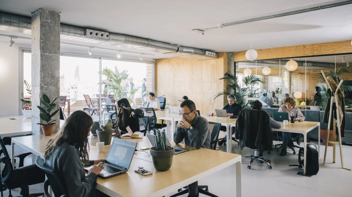 Interior de oficina luminosa con mesas compartidas y luz natural en Cloudworks, fomentando la productividad y el bienestar del equipo.