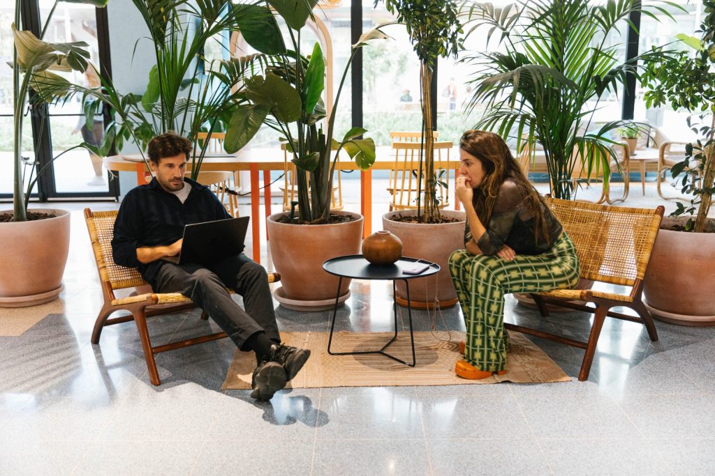 Dos personas en una zona de descanso o "tercer espacio" de una oficina, sentadas en sillas de ratán rodeadas de plantas, fomentando la colaboración informal.