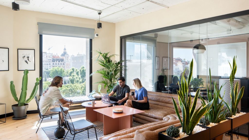 Zona de estar luminosa con sofás de cuero y vistas urbanas, utilizada por empleados para reuniones informales y trabajo relajado en una oficina flexible.